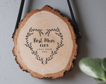 May include: A wooden heart-shaped sign with the text "Best Mum ever" and "Love from Eve" hanging from a black leather strap. The sign is made from a natural wood slice with a rustic finish.