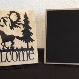 May include: Two square decorative signs. One features a black silhouette of a horse, trees, and mountains with the word "WELCOME" on a beige background. The other sign is a black square with a light-colored border.