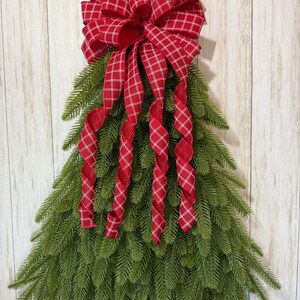 May include: A red and white plaid ribbon bow with tails is attached to a green artificial Christmas tree swag. The swag is shaped like a triangle and is hanging on a white wood background.