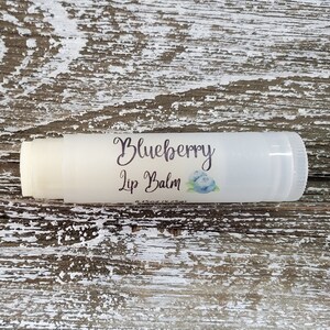 May include: A white lip balm tube with the words "Blueberry Lip Balm" in cursive script and a small blueberry illustration. The cap is off, revealing the balm. The background is a weathered, white-washed wooden surface.