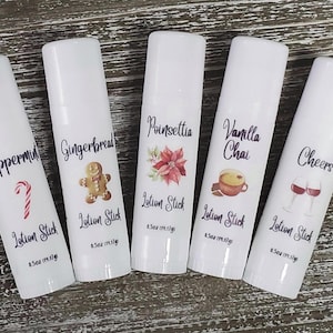 May include: Five white lotion sticks with holiday-themed labels. Flavors include Peppermint, Gingerbread, Poinsettia, Vanilla Chai, and Cheers. Each stick is labeled with the flavor name and a corresponding graphic, such as a candy cane or gingerbread man.