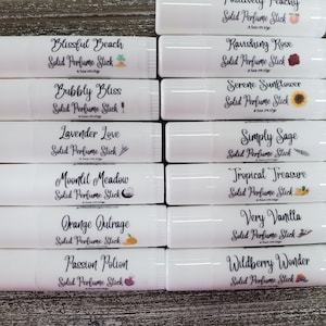 May include: Twelve white tubes with colorful labels and illustrations. Each tube is labeled with a different scent name and the words "Solid Perfume Stick". The scents include Blissful Beach, Bubbly Bliss, Lavender Love, Moonlit Meadow, Orange Outrage, Passion Potion, Positively Peachy, Ravishing Rose, Serene Sunflower, Simply Sage, Tropical Treasure, Very Vanilla, and Wildberry Wonder.