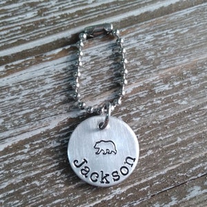 May include: A silver keychain with a round metal tag that says "Jackson" and has a bear silhouette. The keychain has a silver ball chain.