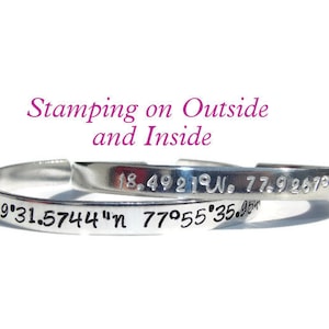 May include: A silver bangle bracelet with coordinates engraved on the outside and inside. The outside reads "18.4921°W. 77.9267°W" and the inside reads "18°29'31.5744"N 77°55'35.95"W".