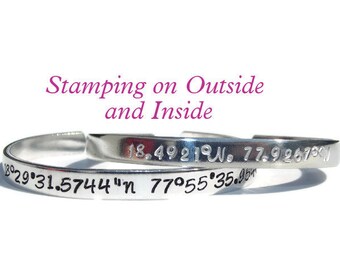 May include: A silver bangle bracelet with coordinates engraved on the outside and inside. The outside reads "18.4921°W. 77.9267°W" and the inside reads "18°29'31.5744"N 77°55'35.95"W".