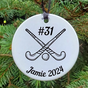 May include: White round ornament with a black hockey stick design, a black ball, and the text "#31 Jamie 2024".
