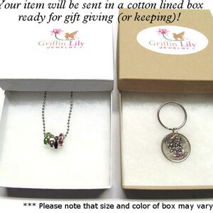 May include: Two gift boxes with white interior and brown exterior. The box on the left contains a silver necklace with a charm made of small colorful beads. The box on the right contains a silver key chain with a round charm that says "Mommy's Best Gift Ever". Both boxes have a Griffin Lily Jewelry logo.