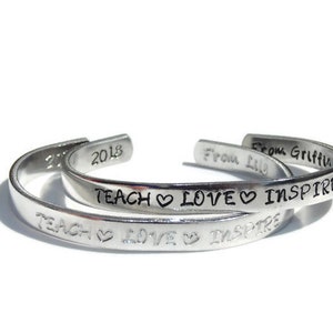 May include: Two silver bracelets with the inscription "Teach Love Inspire" and the year "2018". The bracelet on top also has the inscription "From Griffin".