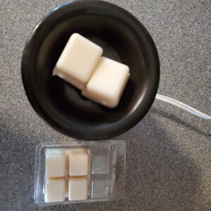 May include: A black wax warmer with two white wax melts inside. A clear plastic package with four additional white wax melts is shown below the warmer.