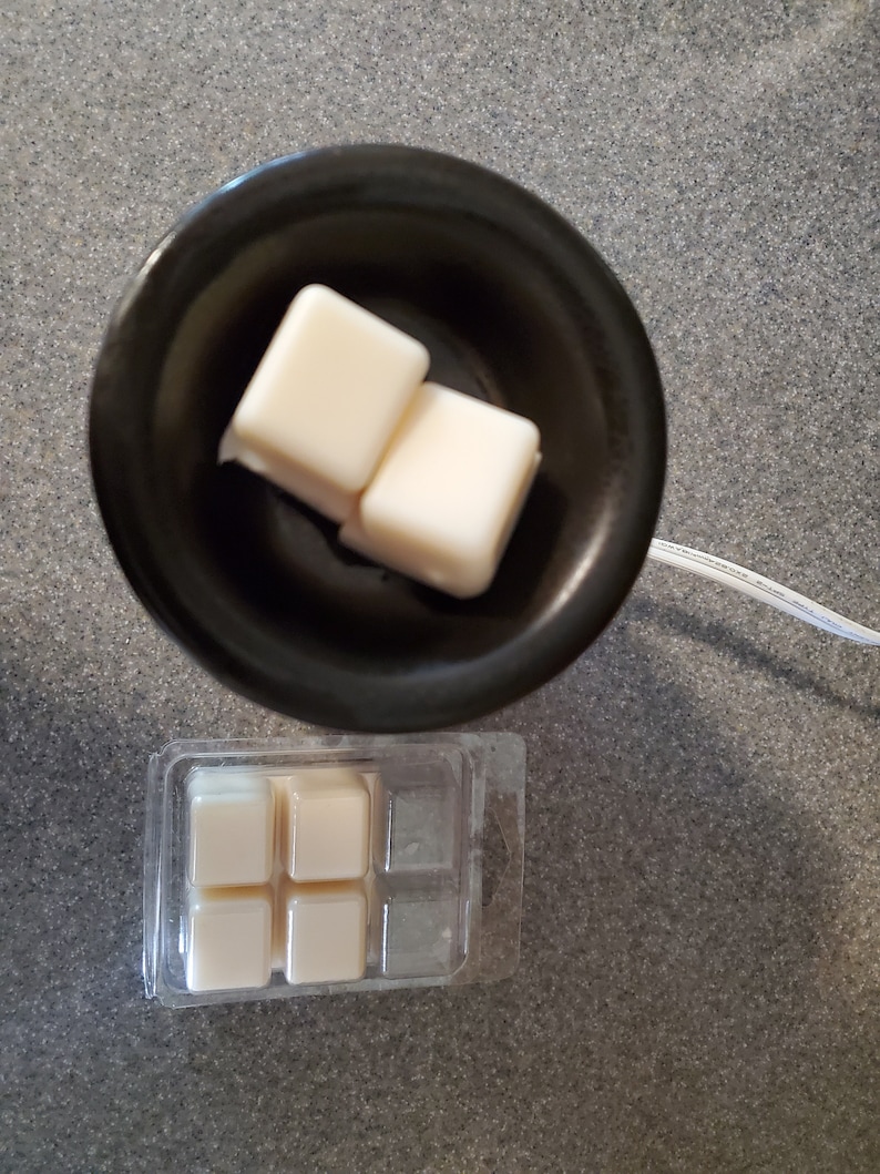 May include: A black wax warmer with two white wax melts inside. A clear plastic package with four additional white wax melts is shown below the warmer.