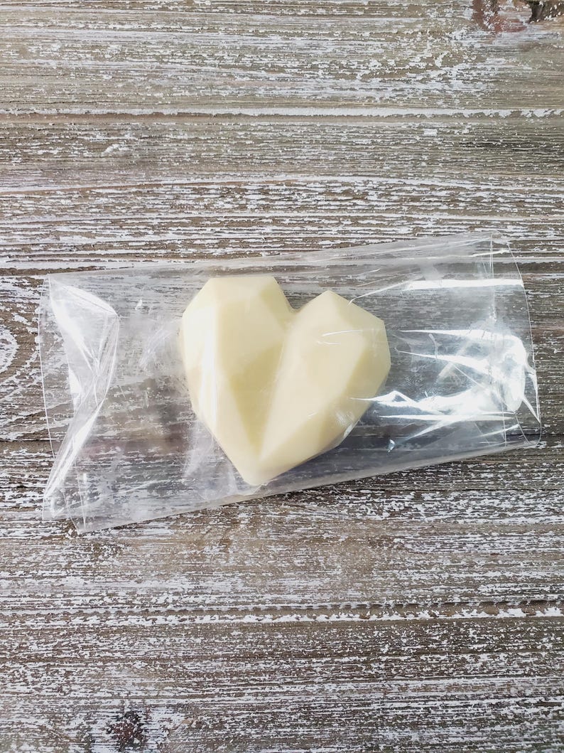 Pode incluir: Um cora&ccedil;&atilde;o de cera branco-sujo com um design geom&eacute;trico est&aacute; dentro de um saco pl&aacute;stico transparente. Est&aacute; posicionado sobre uma superf&iacute;cie de madeira clara e envelhecida.