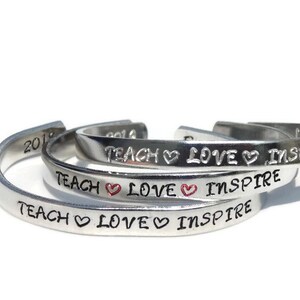 May include: Three silver bracelets with the words "Teach Love Inspire" engraved on each. The word "Love" has a small red heart symbol between the words.