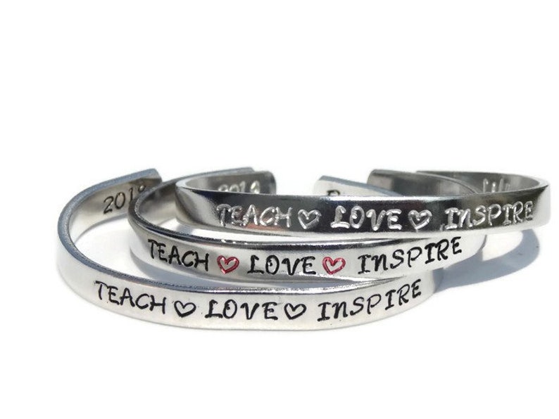 May include: Three silver bracelets with the words "Teach Love Inspire" engraved on each. The word "Love" has a small red heart symbol between the words.