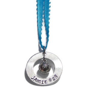 May include: A silver-toned circular metal pendant featuring the engraved text JAMIE #23, with a small three-dimensional basketball charm suspended in the center. The pendant is attached to a bright blue ribbon for hanging as an ornament.