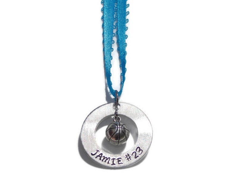 May include: A silver-toned circular metal pendant featuring the engraved text JAMIE #23, with a small three-dimensional basketball charm suspended in the center. The pendant is attached to a bright blue ribbon for hanging as an ornament.