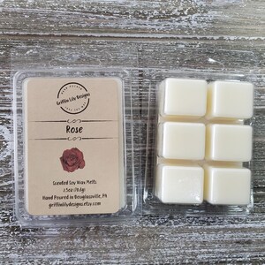 May include: A clear plastic container holding six square wax melts and a label. The label reads "Rose" with a rose graphic and product details. The wax melts are a creamy white color. The product is 2.5oz (70.8g).
