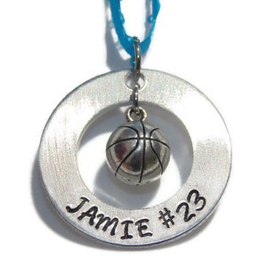 May include: A circular silver-toned metal pendant featuring a brushed finish with the engraved text JAMIE #23. A small, three-dimensional silver-toned basketball charm hangs from the center of the ring, suspended by a bright blue ribbon.