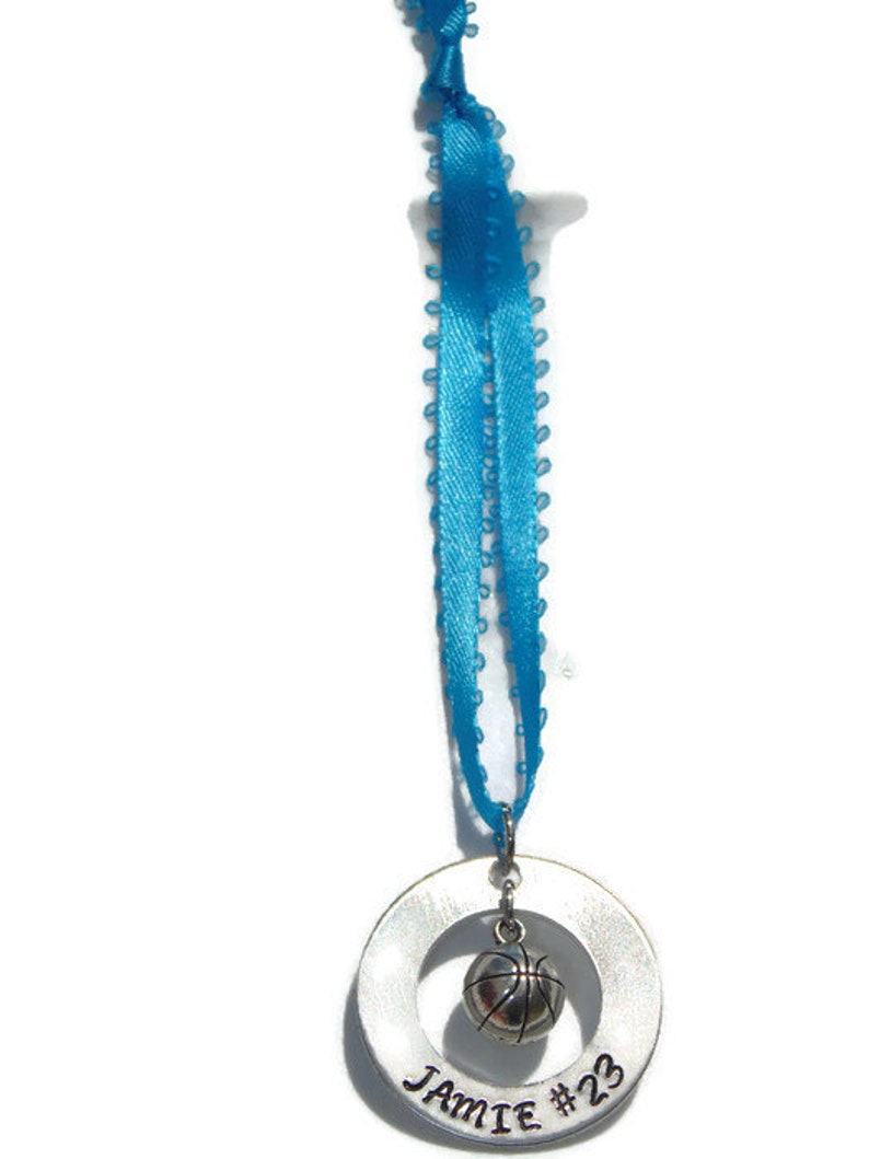 May include: A silver-toned circular metal charm engraved with the name Jamie and the number 23, featuring a small three-dimensional basketball charm suspended in the center, all attached to a bright blue ribbon with a decorative picot edge.