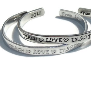 May include: Two silver bracelets with the words "TEACH LOVE INSPIRE" engraved on them. The bracelets are stacked on top of each other. The top bracelet has the year "2018" engraved on it. The bottom bracelet has the words "From As" and "From Gritt" engraved on it.
