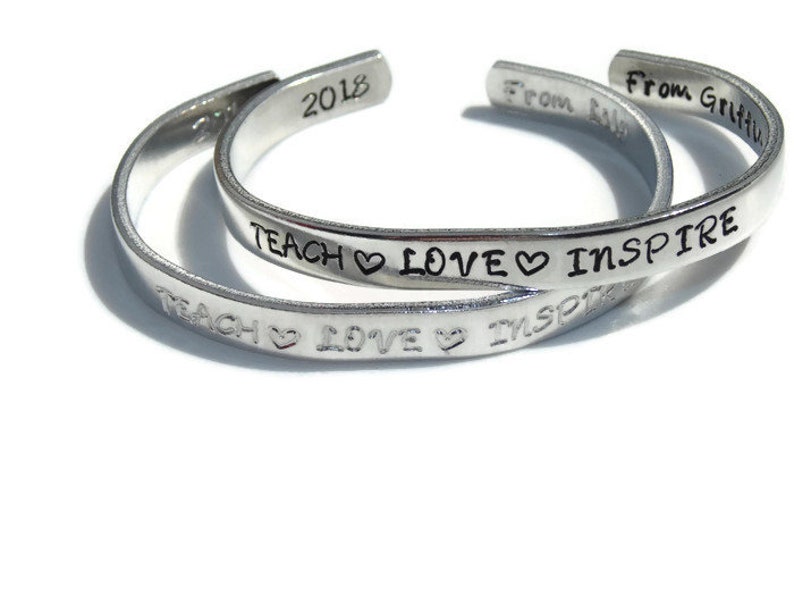 May include: Two silver bracelets with the words "TEACH LOVE INSPIRE" engraved on them. The bracelets are stacked on top of each other. The top bracelet has the year "2018" engraved on it. The bottom bracelet has the words "From As" and "From Gritt" engraved on it.