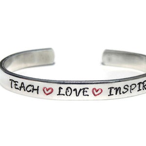 May include: Silver bangle bracelet with the words "Teach Love Inspire" and two red hearts.