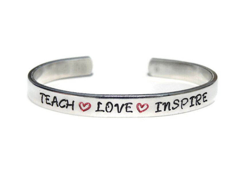 May include: Silver bangle bracelet with the words "Teach Love Inspire" and two red hearts.