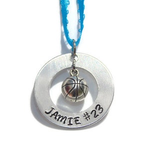 May include: A silver-toned metal circular pendant featuring the stamped text JAMIE #23. A small, three-dimensional silver-toned basketball charm hangs from the center of the ring. The pendant is suspended by a bright blue ribbon loop.