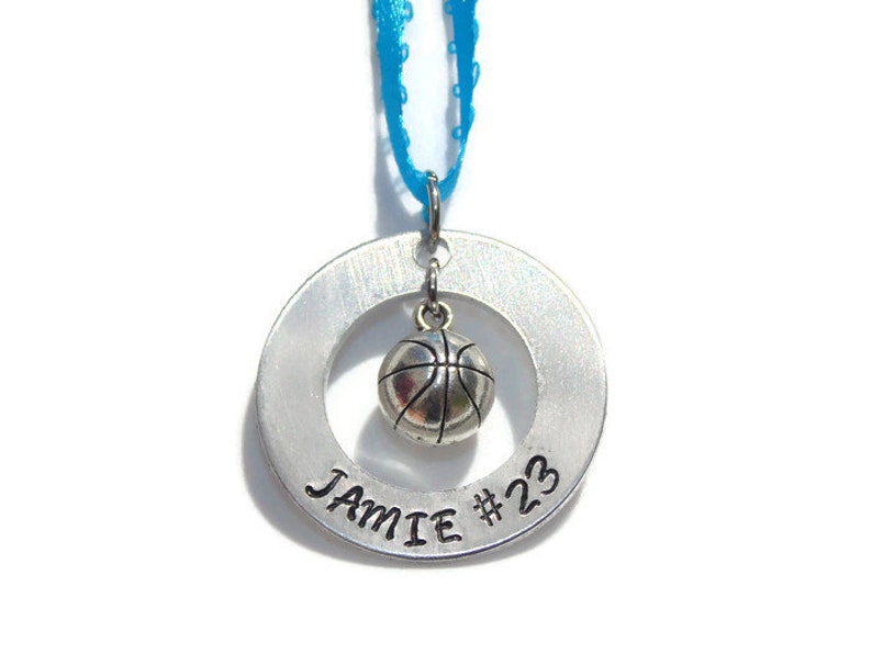 May include: A silver-toned metal circular pendant featuring the stamped text JAMIE #23. A small, three-dimensional silver-toned basketball charm hangs from the center of the ring. The pendant is suspended by a bright blue ribbon loop.