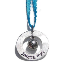 May include: A silver-toned circular metal pendant featuring the engraved text JAMIE #23. A small silver-toned basketball charm hangs from the center of the ring. The pendant is suspended by a bright blue ribbon with a decorative scalloped edge.
