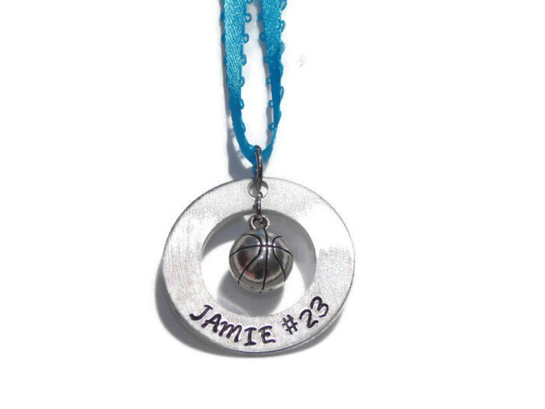 May include: A silver-toned circular metal pendant featuring the engraved text JAMIE #23. A small silver-toned basketball charm hangs from the center of the ring. The pendant is suspended by a bright blue ribbon with a decorative scalloped edge.
