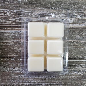 May include: A clear plastic clamshell package containing six ivory-colored wax melt cubes. The cubes are square and evenly spaced within the package. The background is a weathered, gray-toned wooden surface.
