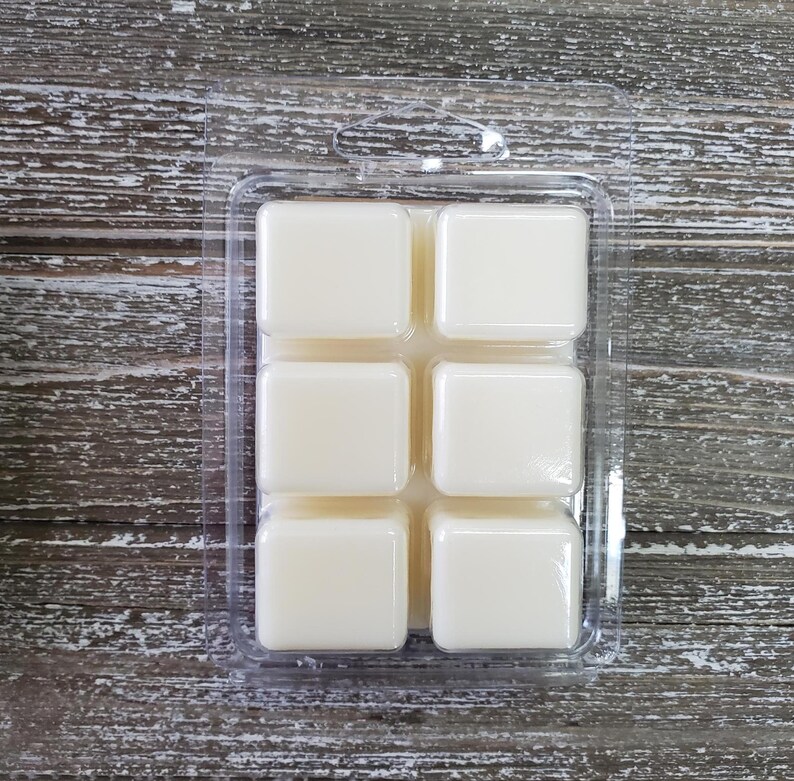 May include: A clear plastic clamshell package containing six ivory-colored wax melt cubes. The cubes are square and evenly spaced within the package. The background is a weathered, gray-toned wooden surface.