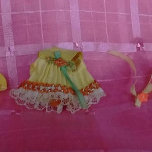 May include: A yellow doll dress with a pleated bodice, a white lace hem, and orange floral accents, accompanied by a matching yellow plastic accessory and a thin yellow ribbon tie with a small orange flower attached to the end of the ribbon.