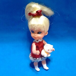 May include: A small doll with blonde hair styled in a high ponytail with a red bow, wearing a sleeveless red dress with silver trim and white shoes. The doll is holding a small white cat figurine with a yellow collar in its arms.