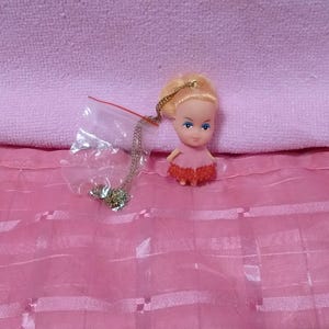 May include: A small doll with blonde hair and blue eyes, wearing a pink dress with a red lace trim. A silver-colored necklace is in a plastic bag. The doll is on a pink surface.