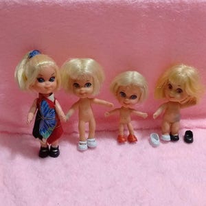 May include: Four small dolls with blonde hair and large eyes. The first doll wears a red dress with a blue butterfly print and black shoes. The second doll is nude with white shoes. The third doll is nude with red shoes. The fourth doll is nude.