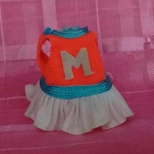 May include: A small sleeveless garment featuring an orange bodice with a light gray letter M centered on the front. The neckline and waist are trimmed with a bright teal braided cord. The bottom consists of a pleated white skirt section.
