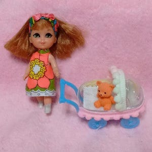 May include: A small doll with auburn hair, wearing a pink and green floral dress and a matching headband, stands next to a miniature blue and pink toy stroller containing a small orange teddy bear with a white blanket inside the carriage.
