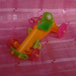 May include: A plastic toy vehicle featuring an orange body, a bright green seat, and a pink steering wheel. The toy has four pink wheels, a green front bumper, and a small green pedal mechanism located near the center of the orange frame.