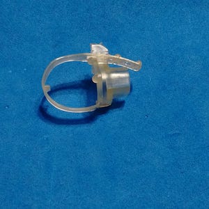 May include: A clear, translucent plastic ring component featuring a small, cylindrical protrusion and a thin, flexible strap with a series of small holes, designed for securing or fastening purposes in a mechanical or assembly application.