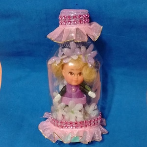 May include: A clear plastic bottle decorated with pink and white accents, containing a small doll. The bottle is adorned with pink fabric, rhinestones, and floral embellishments. The doll wears a purple dress and has blonde hair.