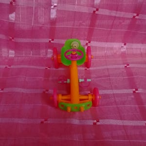 May include: A colorful toy car with an orange body, green seat and wheels, and a pink steering wheel. The toy car has a small doll figure in the seat. The toy car is on a pink background.