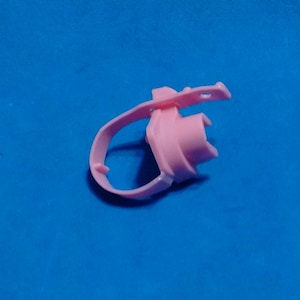 May include: A light pink plastic ring-shaped component featuring a central cylindrical housing and an attached flexible strap with a small circular hole at the end. The object has a smooth, matte finish and a uniform pastel pink color throughout.