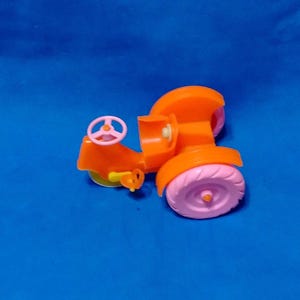 May include: An orange toy road roller with pink wheels and a pink steering wheel. The toy has a small yellow roller and is set against a blue background.