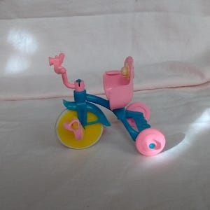 May include: A small, colorful toy tricycle in pink, blue, and yellow. The tricycle has a pink seat and handlebars, blue frame, and yellow wheels. The wheels are round and the seat has a small backrest.