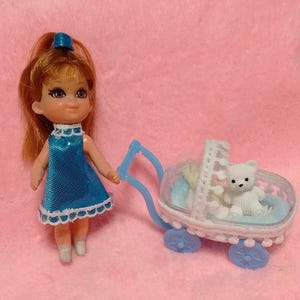 May include: A doll with auburn hair tied with a blue bow, wearing a shiny blue dress with white scalloped trim. Beside the doll is a light blue plastic toy stroller with a white lace-like canopy, containing a small white bear figurine inside.