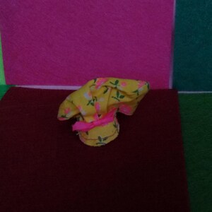 May include: A small, yellow chef's hat with a floral pattern in pink and green. The hat is tied with a pink ribbon. The hat is sitting on a dark red surface.
