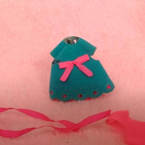 May include: A teal felt dress with a pink bow and scalloped hem. A pink ribbon and a detached pink felt piece are also present. The items are on a pink, textured surface.