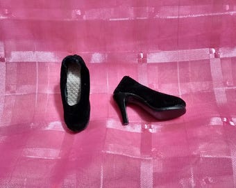 May include: Two black doll shoes are displayed on a pink surface. One is a flat shoe, and the other is a high-heeled pump. The shoes are made of a soft, velvety material.