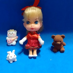 May include: A small doll with blonde hair, a red bow, and a red dress, accompanied by a white bear figurine holding a pink guitar, a small white rabbit figurine, and a textured brown teddy bear figurine, all arranged together as a set.